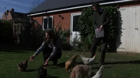 Andrew Davies and his wife Milca in a garden. She is crouching on grass in front of a building feeding chickens, while a he is standing behind her holding a box of chicken feed.