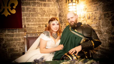 Ian Gilbert Seated portrait of two people in medieval-inspired attire inside a stone-walled room lit by a warm lantern. Jasmine who has ginger hair wears a white flowing dress with decorative silver shoulder pieces and a jewelled headpiece, while her husband wears dark armour with a green sash draped across the chest. They sit closely together on an ornate wooden chair beside a red banner with a gold emblem.