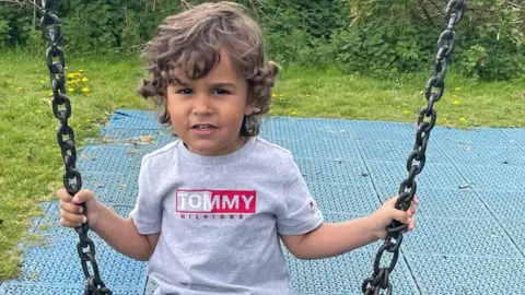 A boy with curly brown hair looking at the camera on a swing.