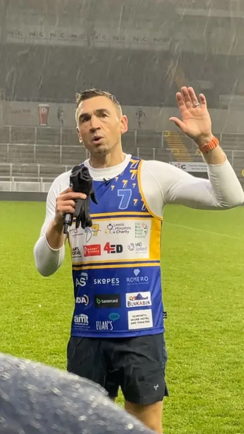 Kevin Sinfield with microphone and gloves in one hand raises his other hand during a speech at Headingley Stadium.