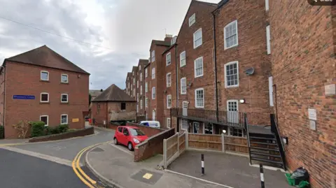 The entrance to Warmstry Court, which is off Quay Road. The apartment complex has large buildings containing flats. A blue sign reads Warmstry Court on one side of the road. There are double yellow lines and a car is parked ina  parking space. Stairs lead up to an entrance and walkway on the side of one of the buildings.