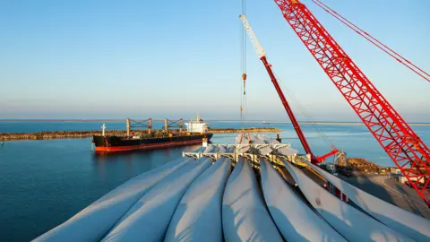 Getty Images A crane lifts large turbine blades towards a boat on the background. There is a calm blue sea and a rock breakwater on the water.