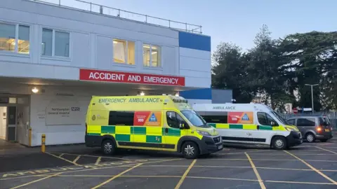 White two-storey emergency department showing exterior and ambulance parking bays - with a yellow and a white ambulance parked. There is a sign over the entrance indicating "Accident and Emergency" and trees to the right.
