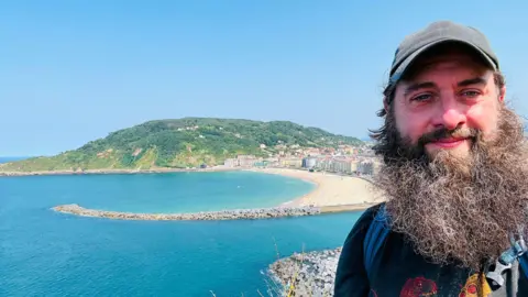Ryan Milligan Man with beard and a cap standing on a higher area with the view of a beach behind him.