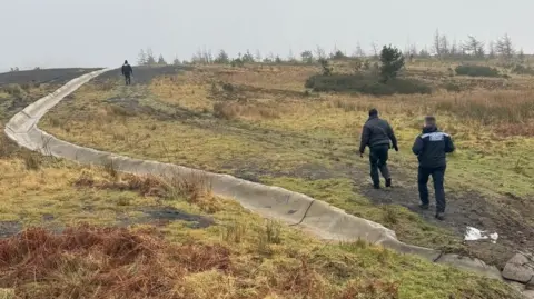 South Wales Police police officers walking on a muddy track beside a drainage channel in a boggy area. 