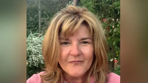 A selfie image of Nichola Greenfield. She has shoulder length hair and is wearing a pink hoody. She is standing in front of some trees. 