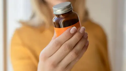 A woman (neck below) in a yellow jumper holding a bottle of medicine with an orange wrapper.