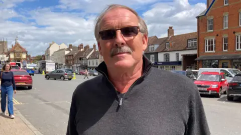 Gary Farrow. He is pictured in Marlborough town centre. He has short grey hair and a grey moustache. He is wearing dark square-shaped sunglasses.