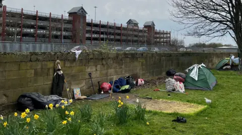 A number of tents can be seen on a thin strip of grass, against a wall; a patch of dead grass outlines where a tent previously sat. Bags of belongings and duvets can be seen strewn around the grassy area, a bare tree stands to the far right.