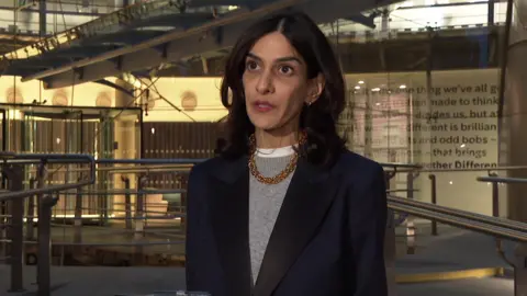 Priya Dogra in dark jacket, grey jumper and gold chain, shoulder length dark hair. Standing in front of steel railings at Channel 4