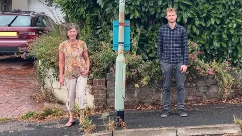 Roberta Ayles on the left in a flowery top and Ethan Merrington-Pink on the right in a check shirt with a bus-stop between them 