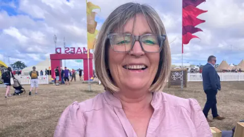 Anna Brychan at the Eisteddfod festival in mauve shirt laughing at the camera. She has a blonde bob and wears glasses. Festival flags are visible in yellow and red in the background and the grass is dry and brown