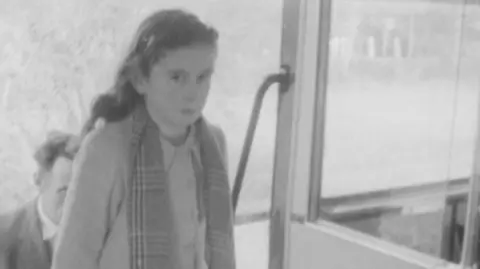A still frame from a black‑and‑white film showing a young girl wearing a coat and patterned scarf standing by the open door of a railbus with another passenger partially visible behind them.”
