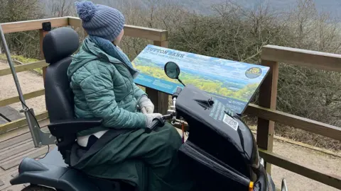 A young woman with a woolly hat sits on a large black off-road mobility scooter. She wears a green puffed jacket and a blue and white striped jumper. She is parked on a raised wooden viewing platform overlooking a countryside scene.