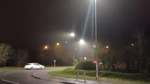 A road junction with street lights and a bus stop. It's dark and a white car is on the road.