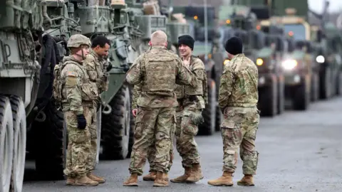 Six US soldiers in green camouflage uniforms are pictured in Germany standing in a circle, with a long line of military vehicles visible behind them.