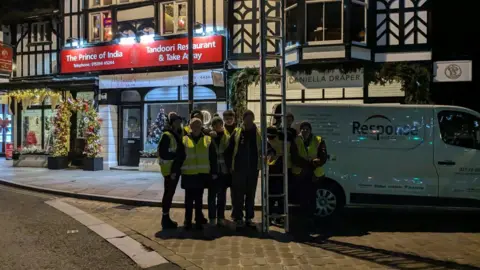 Windermere & Bowness Christmas parade Eight volunteers wearing high-vis vests in Windermere town centre. They are posing for the shot outside an Indian restaurant, holding a tall set of ladders.