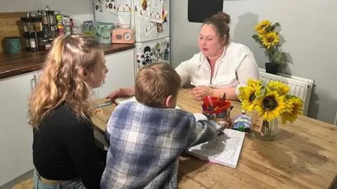 A family of three - a mother with her daughter and son - sat a dining table in their kitchen