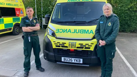 Paramedics Kevin and Amy are wearing all-green uniforms and are standing either side of the front of a yellow ambulance