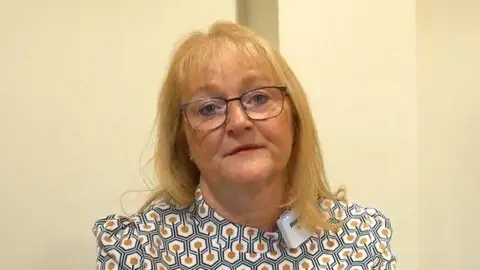 A woman with blonde hair stands against a cream-coloured wall. She is wearing glasses and a hexagonal-patterned blouse. She has a portable lapel microphone on her collar.