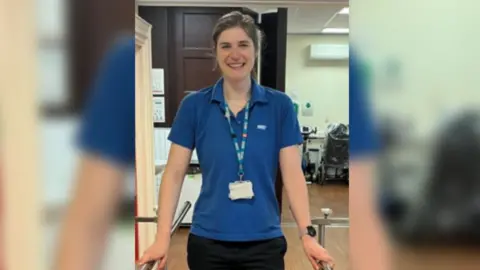 University Hospitals Coventry and Warwickshire A woman wearing a blue NHS top and a lanyard is smiling at the camera. She has dark hair.