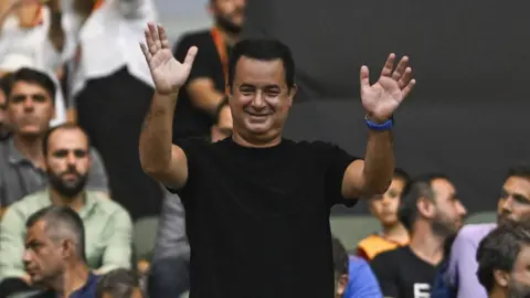 Getty Images A man with short dark hair and wearing a black T-shirt stands up from a crowd of people, waving with both hands in the air.