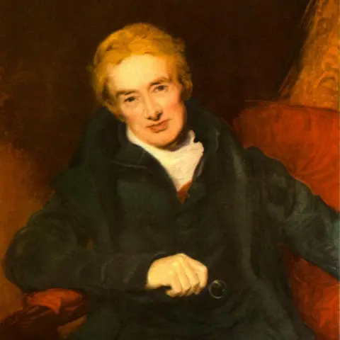 Culture Club/Getty Images An oil painting showing a man with fair hair who is dressed in clothing of the early 19th Century, including a black jacket and white scarf, and smiling as he sits on a red chair.
