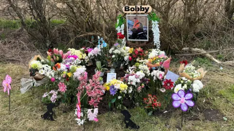 Kimberley Piper/BBC Flowers, photos and tributes left on the side of the road at the site of a traffic collision. 