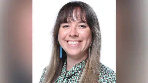 Ffion Mitchell-Langford A woman with long light brown hair, dark brown eyes and fringe is smiling into the camera. She has a green, cream and black chequered shirt on. The background is white with nothing else in frame. She has a long blue decoration on her right ear