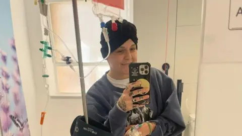 The image shows a woman standing in a hospital room taking a mirror photo while attached to multiple IV lines and a portable infusion pump on a drip stand. The person is wearing casual clothing, including a sweatshirt, joggers and trainers, with medical tubing visible on the arm and wrist. The room appears clinical and brightly lit, with medical equipment and signage visible in the background.