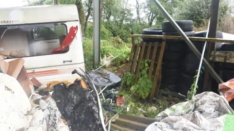 Avon and Somerset Police An abandoned caravan inside a vacant lot. The windows are smashed and there is debris scattered around- including stacked tyres, wooden crates, burnt leather furniture, a corrugated tin roof and an old duvet. The whole site is overgrown and surrounded by trees.