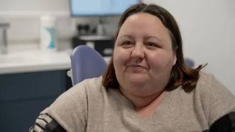 A woman with shoulder-length brown hair and a jumper sits at a dentists.