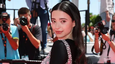 Getty image Mickey Madison at Cannes Film Festival, she is smiling on red carpet and looking at her shoulder in one shoulder, in a black dress 