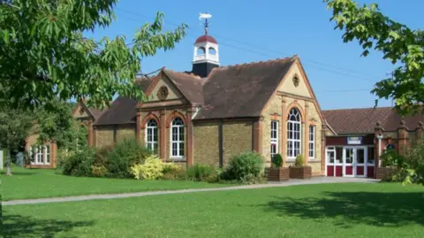 Rob Emms/Geograph The school building set back on a lawn with trees around. It is an older Georgian style building made of yellow-brown brick and with a gable on the roof and arched windows. To one side is a more modern extension with red and white PVC doors.
