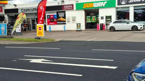 A painted road marking points to a row of shops and a petrol station's no entry junction