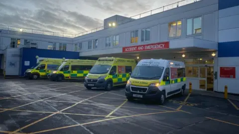 Three yellow ambulances and one white ambulance with green and yellow trim parked on yellow hashed ground in front of a white two-storey Accident and Emergency Department with the department name in white writing on a red sign above the entrance portico.