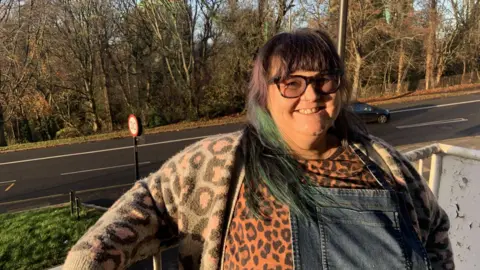 BBC A woman has long black hair with a green streak. She is wearing a patterned cardigan, a leopard print top and dungaree dress and is standing on a council flat veranda with woodland in the background.