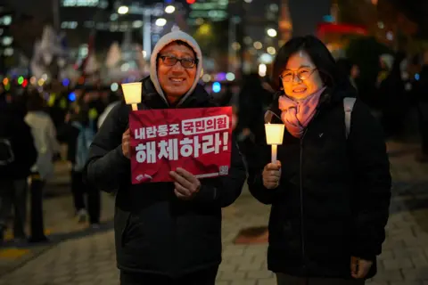 A man and a woman stand at a protest, holding a sign and candles