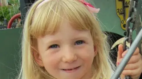 Supplied A young Ava with long, light-blonde hair is pictured outdoors, holding onto a length of chain with one hand. The hair is partially held back with a light pink headband that has a small bow on one side. The background includes part of a large, industrial-looking metal structure painted green, along with a yellow panel featuring dark numbers and markings. Some greenery and buildings can be seen further in the distance.