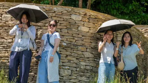 Getty Images Four young women stand by a sandstone wall in the Cotswolds village of Bibury. They are dressed in jeans and light-coloured tops and are sharing two umbrellas between them as they shade themselves from the sun