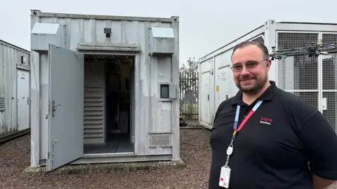 Martin Heath/BBC Alan Storer with short dark hair and beard wearing a black Arqiva T-shirt and a red lanyard standing outside a square grey metal cabin with its door open. An equipment rack can be seen inside. There are more grey cabins on either side, which can be seen to extend some distance from the door.