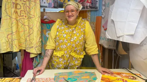 Zoe Dennis/BBC May Gauntlett leaning on her work table with a pair of fabric scissors under her right hand. She is wearing a variety of yellow, patterned clothing, including a headscarf. Fabric and design images are laid on the table and hanging behind her