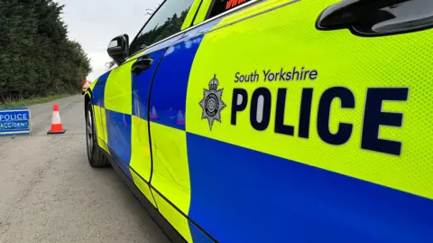 A South Yorkshire Police car with bright yellow‑and‑blue markings parked on a road, with a traffic cone and a blue “Police Accident” sign set up nearby, against a backdrop of trees.