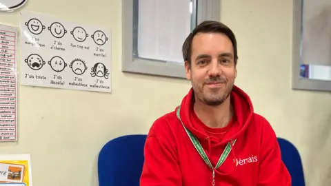 Ben is wearing a red hoodie and sitting on a blue chair in a classroom, with French-language posters, charts, and books visible on the walls and shelves behind him.