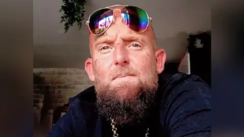 Lancashire Police A selfie of Anthony. He is a white man with a medium-length brown beard and bald head. He is wearing a navy blue t-shirt, gold chain and rainbow-lensed aviator sunglasses.
