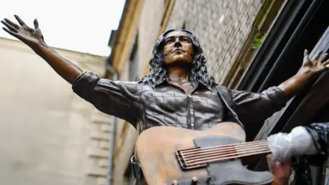 Pacemaker Bronze statue of Rory Gallagher with a guitar hanging off him, photographed against brick wall at Ulster Hall