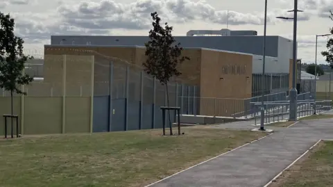 A wired fence leads around the prison and up to a brown bricked building with a sign on the front. A concrete walk way leads to the prison's entrance.