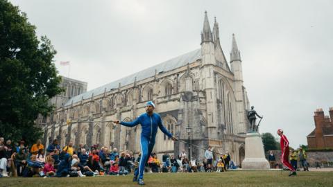 Two men performing in front of crowds outside Winchester Cathedral. The men are in blue and red outfits and holding poles, which looks as though they are juggling.
