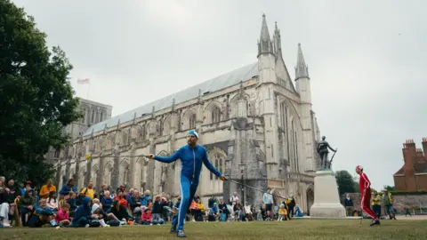 Adrienne Photography / Hat Fair Two men performing in front of crowds outside Winchester Cathedral. The men are in blue and red outfits and holding poles, which looks as though they are juggling.