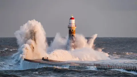 Roker Pier. Large waves are crashing into the pier and it's lighthouse. The pier curves to the right and has railings along each side. The lighthouse has a white and red dome.